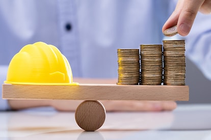 A wooden scale with balancing coins and a yellow hard hat