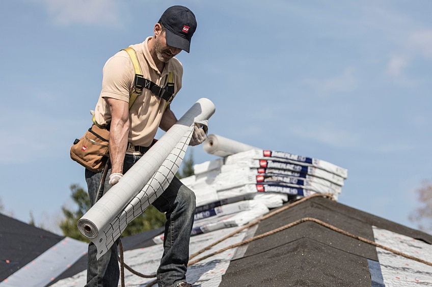 A worker uses thick boots to unfurl a roll of roofing felt while standing