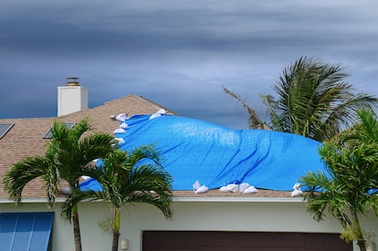Storm damaged roof on house with a protective blue plastic tarp spread over hole in the shingles and