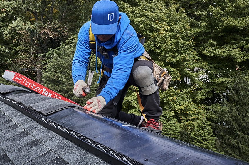 Roofer installing GAF TimberTex ridge cap shingles on the roof of a house.