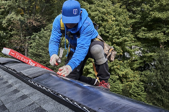 Roofer installing GAF TimberTex ridge cap shingles on the roof of a house.