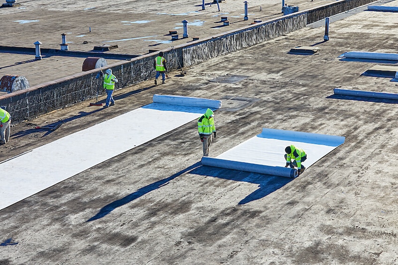 A worker lays out a roll of TPO roofing membrane on a rooftop.