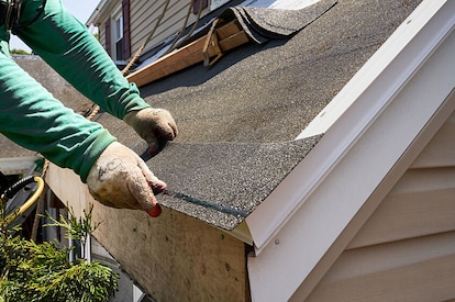 A roofer stands on a ladder and repairs a home's soffit and fascia.