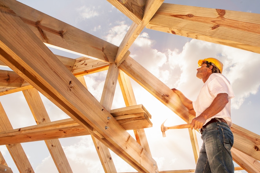 A roofer on rafters hammers a nail.