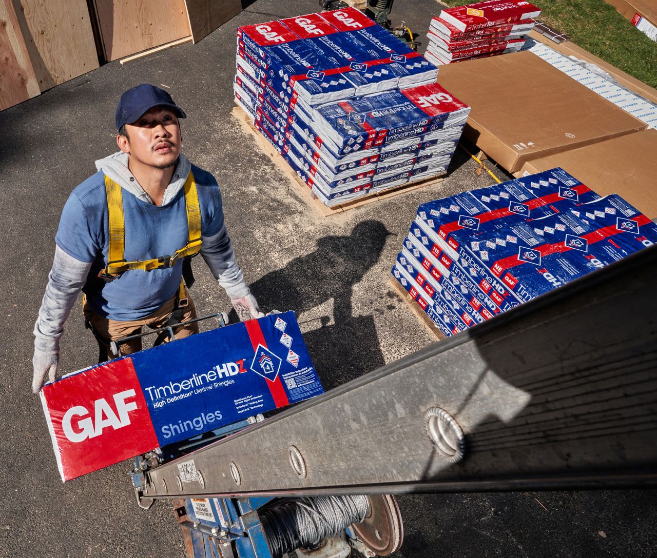 c9d7f984-a997-4f61-8f52-949f6ddca396 A roofer carries a bundle of GAF shingles up a ladder onto a roof.