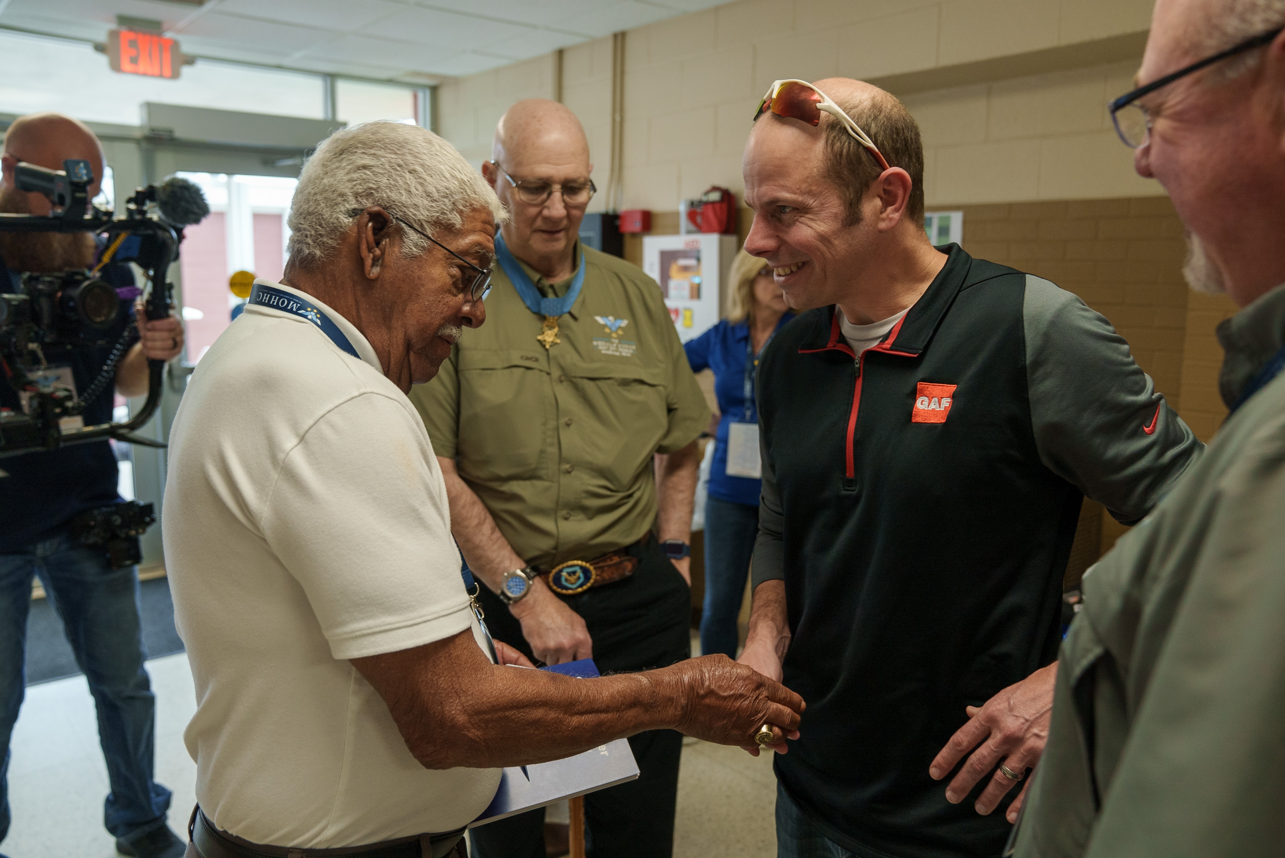 A smiling white man with brown hair shakes hands with a man with brown skin and grey hair while receiving a medal of honor coin. A medal of honor recipient stands looking on in the background.